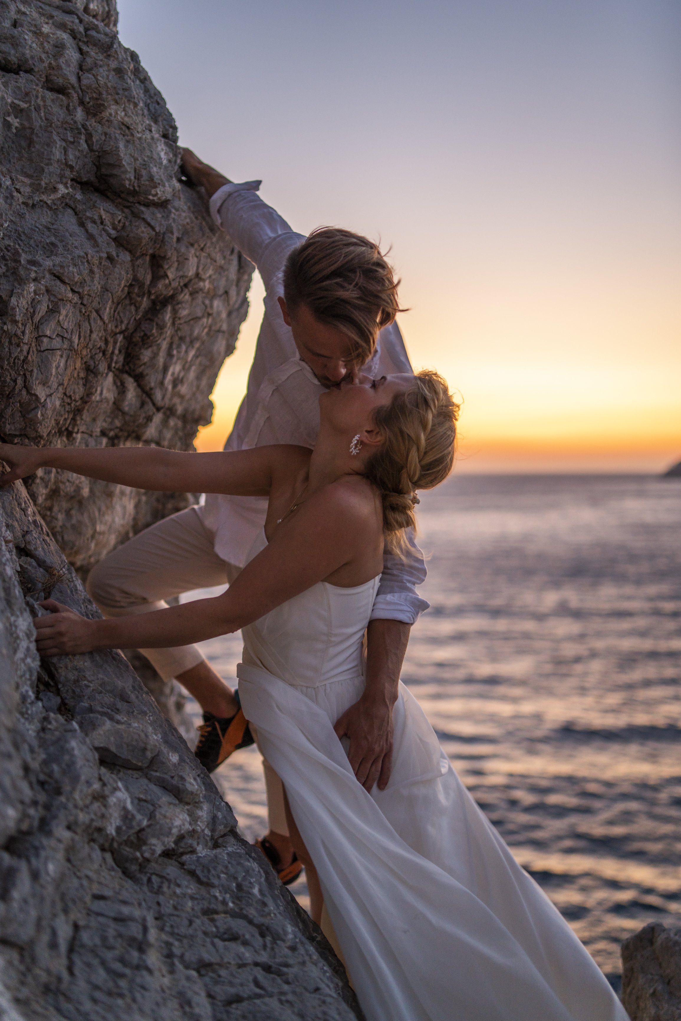 A couple sharing a kiss on a rocky beach at sunset, with the ocean in the background.