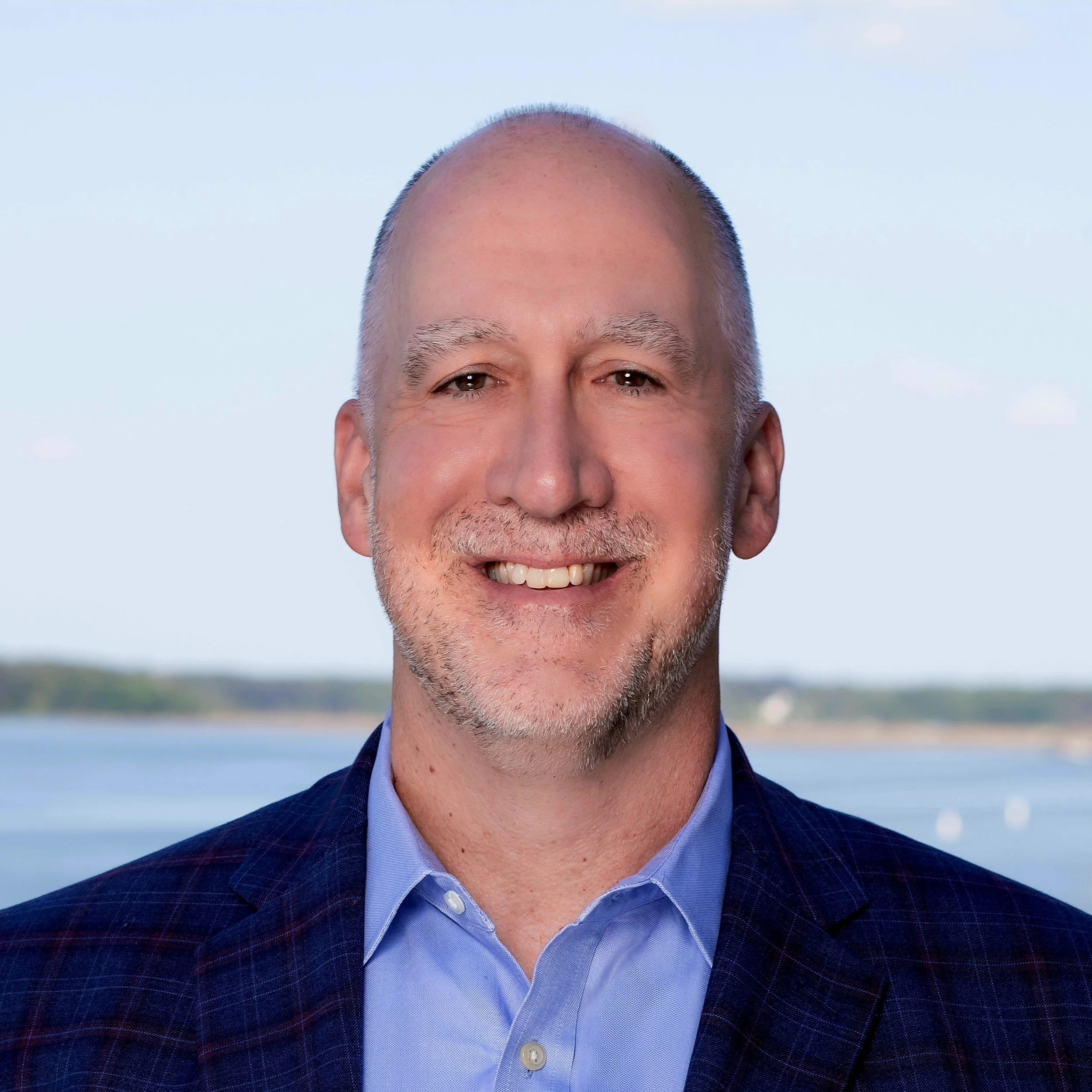 Portrait of a smiling middle-aged man with short gray hair and a beard, wearing a blue blazer and a light blue shirt, outdoors near water with a clear sky in the background.