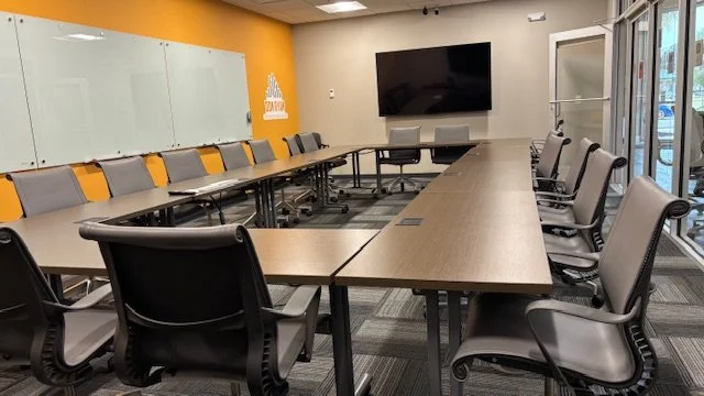 Conference room with U-shaped table, black office chairs, a large TV screen, a whiteboard on the wall, and a large window.
