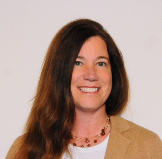 A woman with long brown hair smiling, wearing a beige blazer and a necklace, against a plain white background.