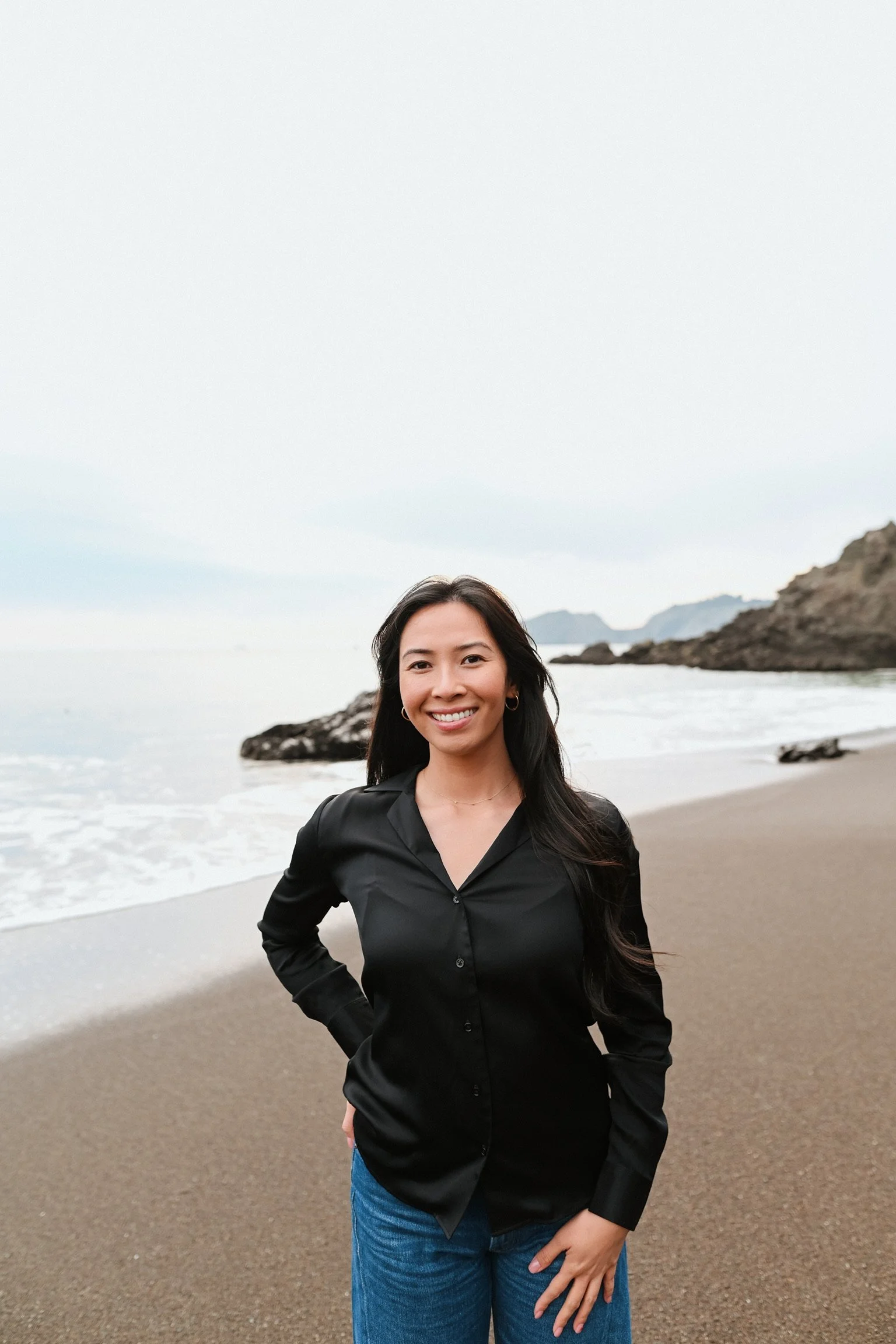 A woman standing smiling, with long dark hair, wearing a black shirt and blue jeans.