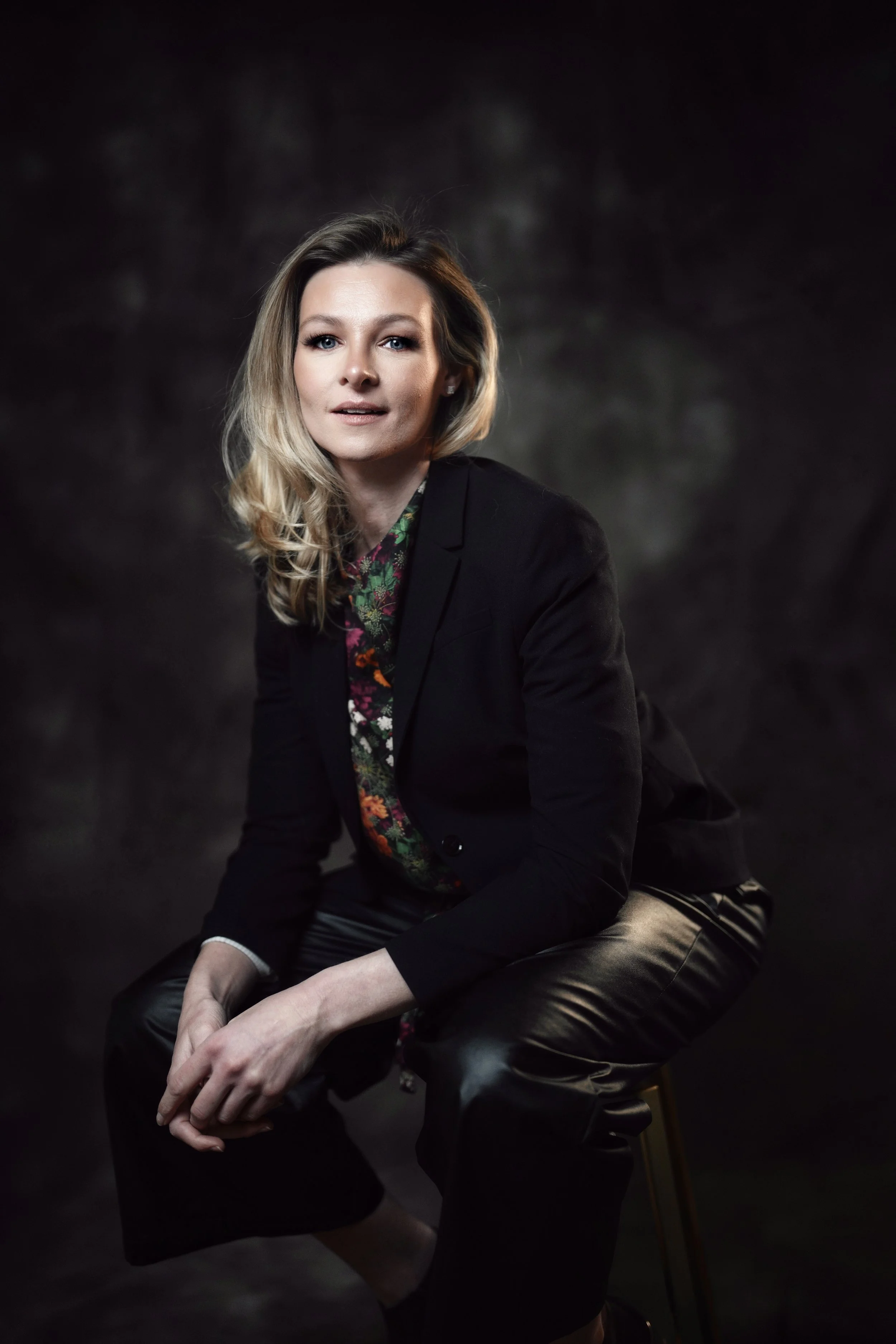 A woman with wavy blonde hair, wearing a black blazer and floral blouse, sitting on a chair in front of a dark, textured background.
