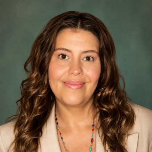 A woman with wavy brown hair, wearing a beige jacket over a blue chambray shirt, and a colorful beaded necklace, smiling against a plain light background.