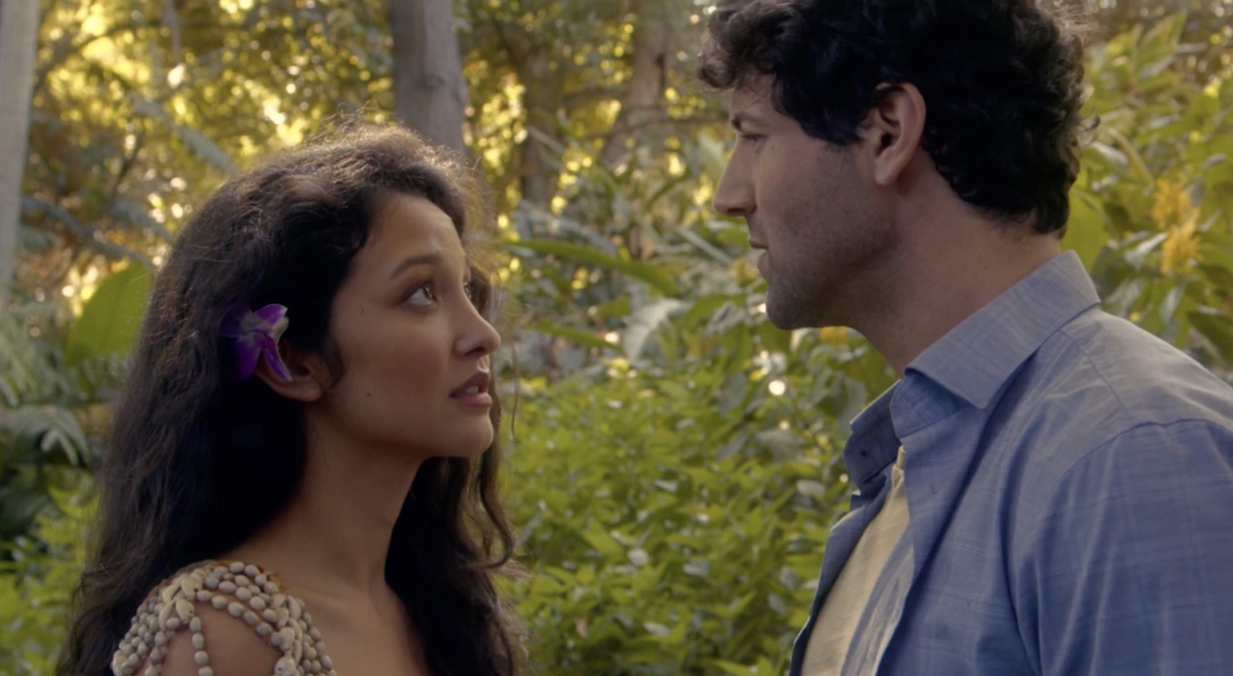 A woman with long dark hair and a purple flower in her hair looks into the eyes of a man with curly dark hair, against a background of green trees and sunlight filtering through the leaves.