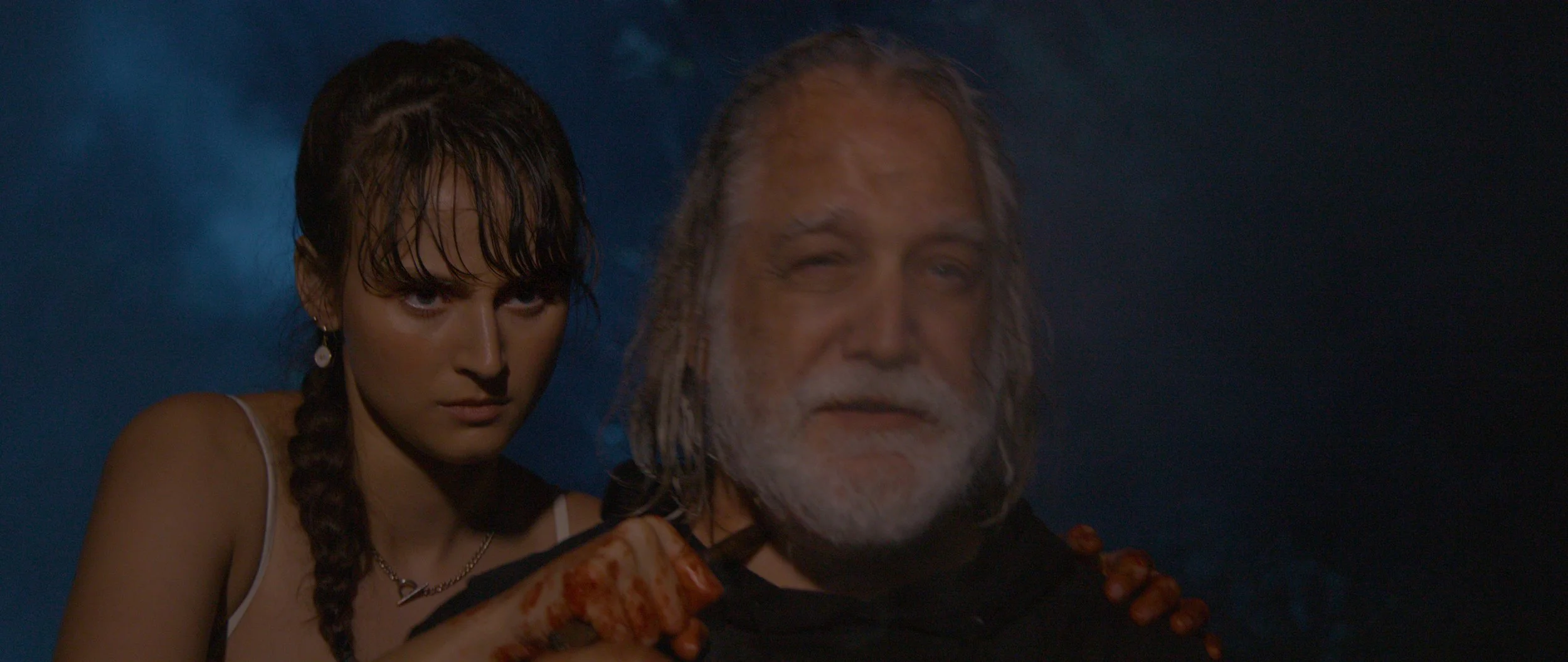 A woman with wet hair and braided pigtails standing behind an older man with a beard, both looking serious and intense, with a dark and smoky background.