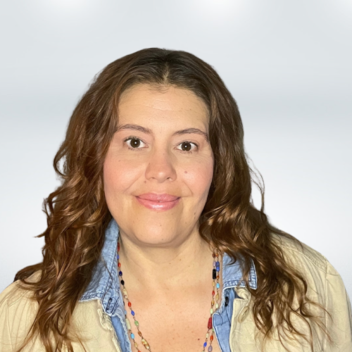 A woman with wavy brown hair and light skin, wearing a denim shirt and a colorful beaded necklace, smiling at the camera against a light background.