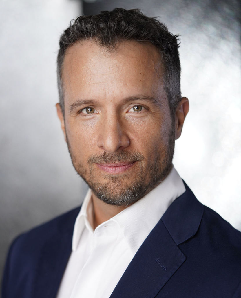 Headshot of a man with short dark hair, some gray, and facial hair, wearing a white shirt and navy blazer, looking at the camera with a confident expression.