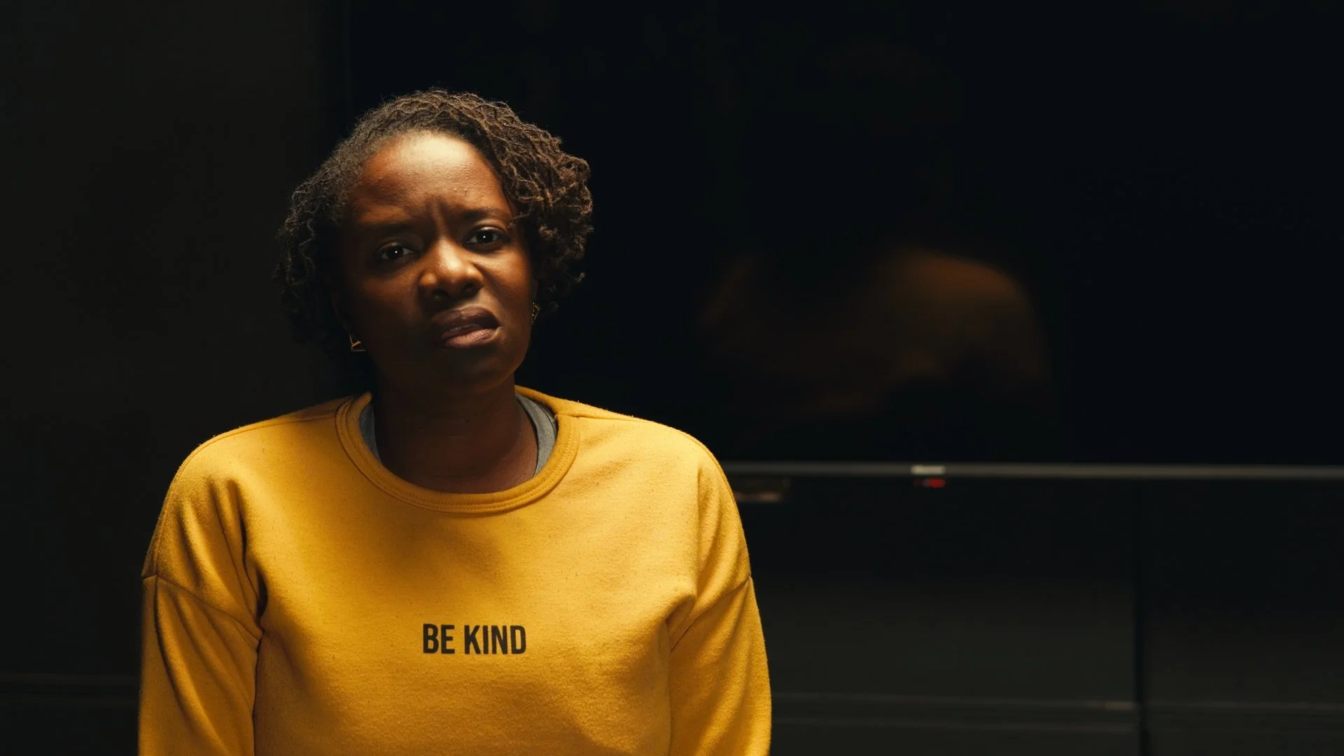 A woman with short, curly hair wears a yellow sweatshirt with the words 'BE KIND' printed on it, standing against a dark background with a large black screen or monitor behind her.