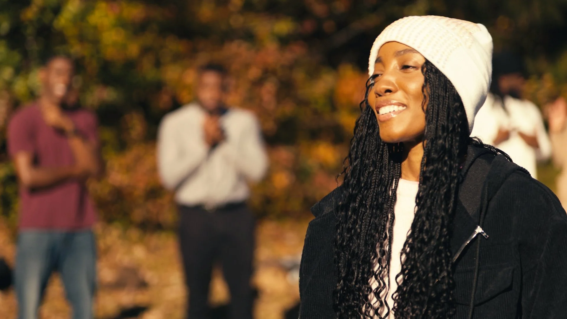Smiling woman with long curly hair wearing a white knit beanie and black jacket, standing outdoors with fall foliage, with blurred group of friends in the background.