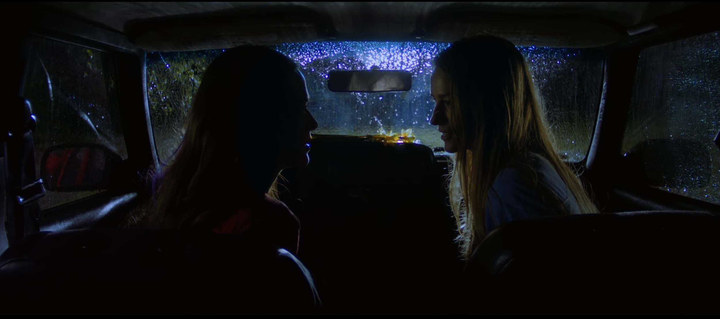 Two women sitting inside a vehicle at night with rain on the windows, facing each other and smiling, illuminated by blue and purple fairy lights outside.