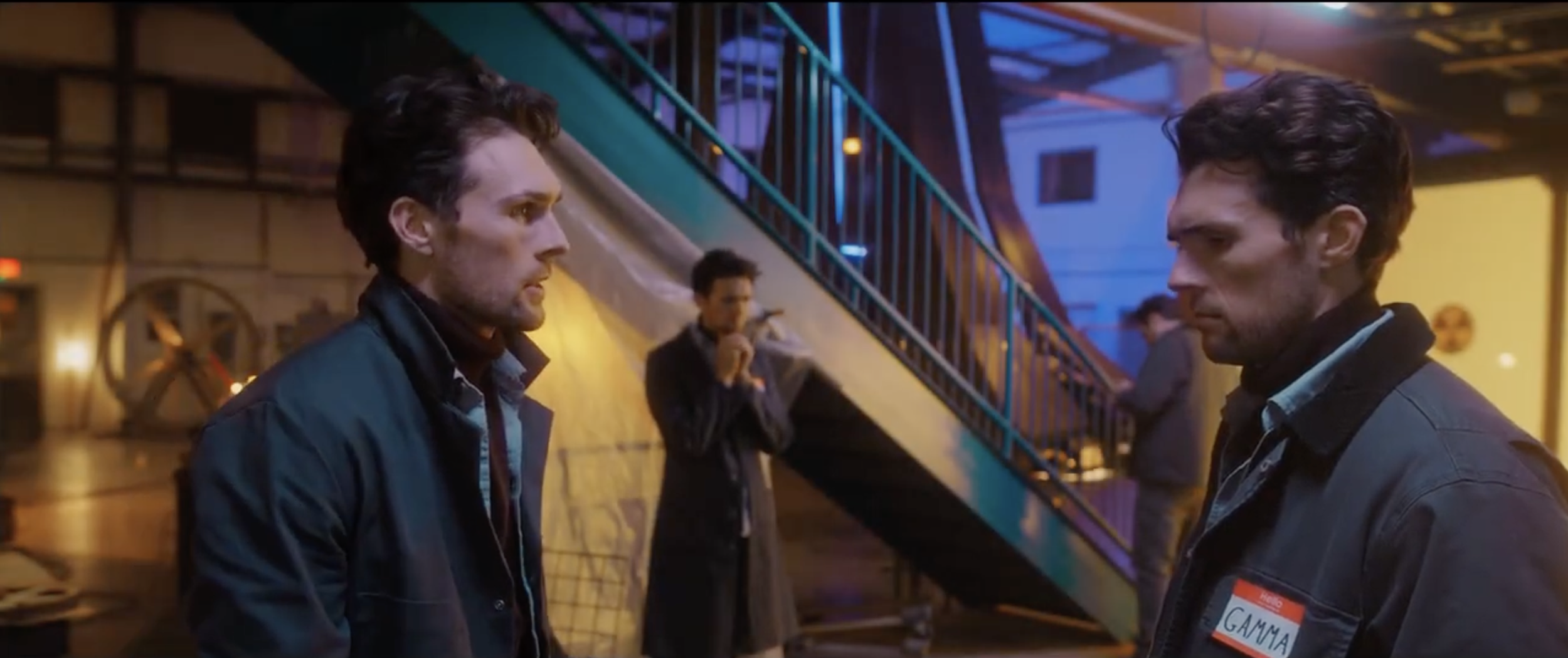 Two men with dark hair face each other in a discussion, inside an indoor amusement park or arcade with colorful lighting and a staircase in the background.