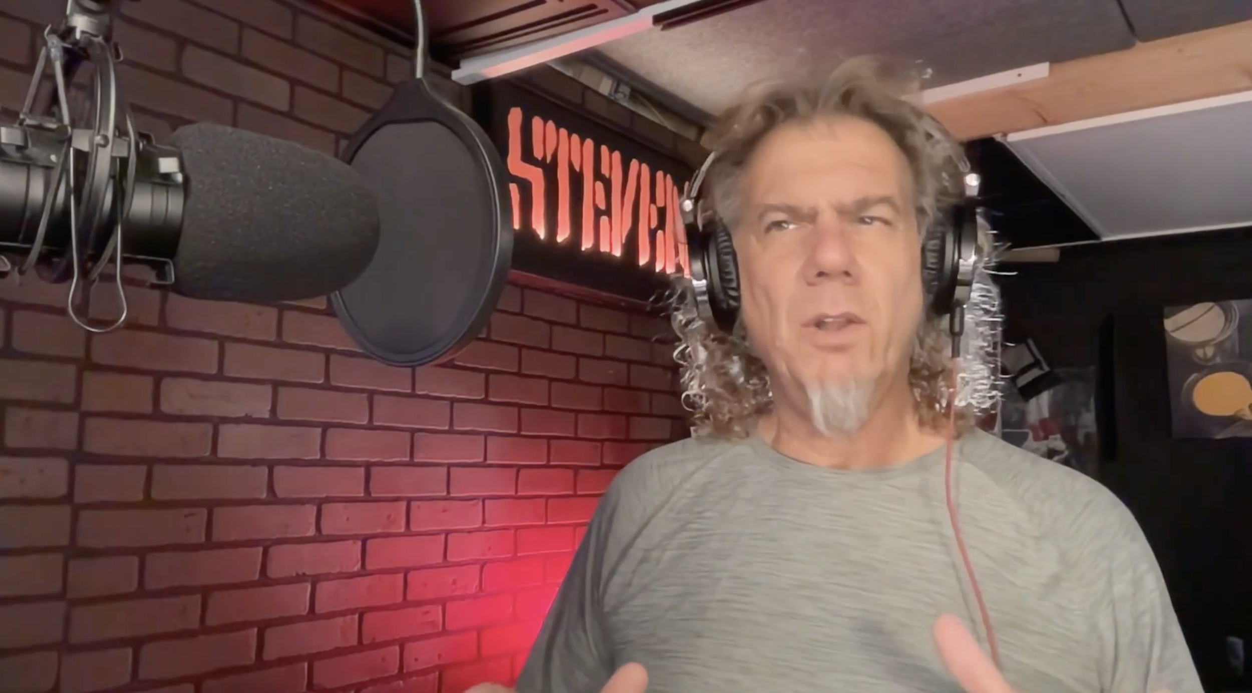 A man with curly gray hair and a beard wearing headphones speaking into a microphone, with a red brick wall and a neon sign in the background.