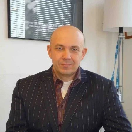 Bald man in pinstripe suit sitting in office with window blinds and a lamp in the background.