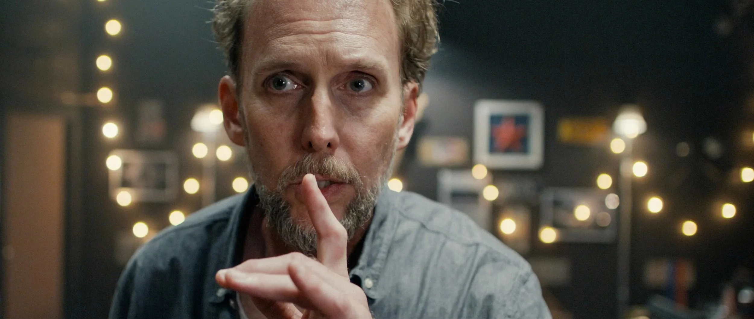 A man with gray hair and beard making a 'shushing' gesture with his finger on his lips, in a room with blurred string lights and framed pictures in the background.