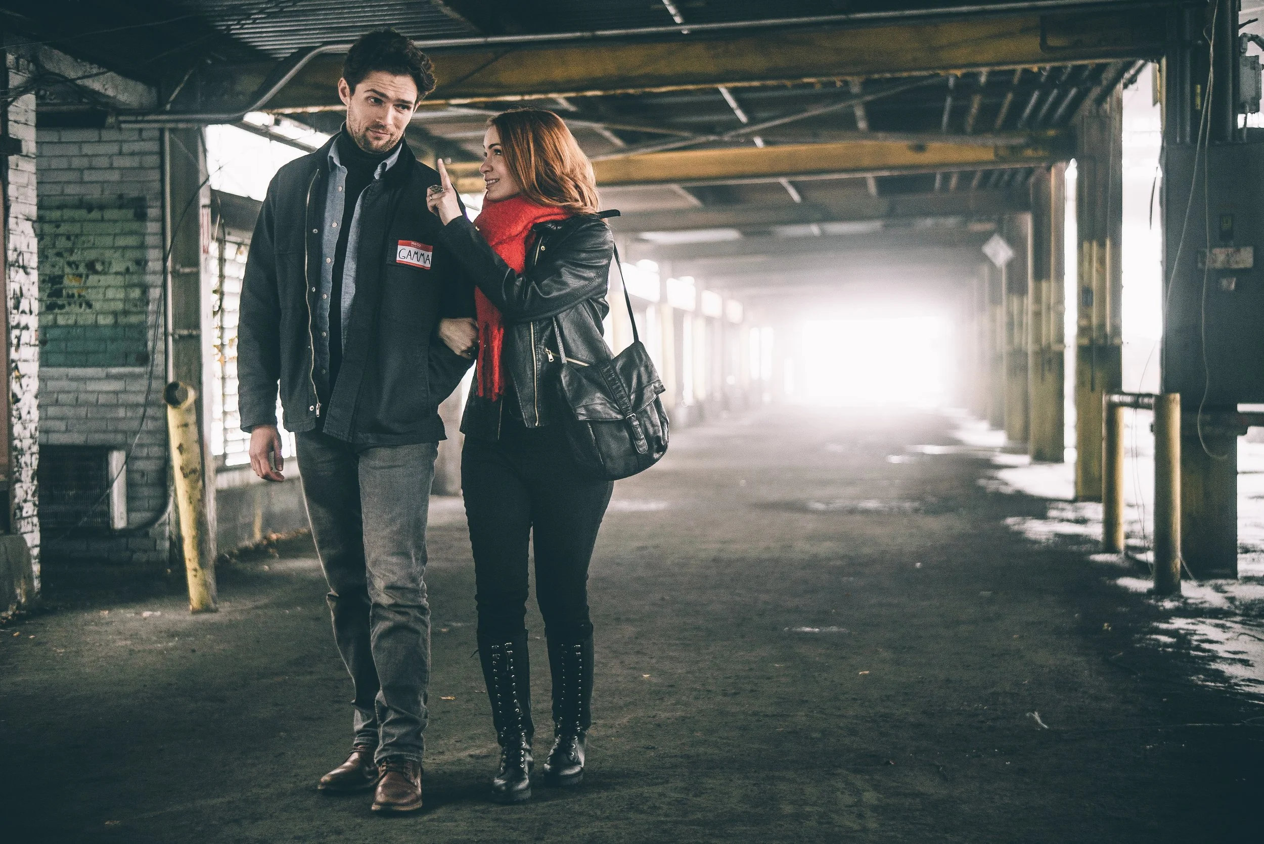 A man and a woman walking together in an urban, underpass-like setting with exposed wooden beams and brick walls. The woman is pointing her finger at the man's chest while they talk.
