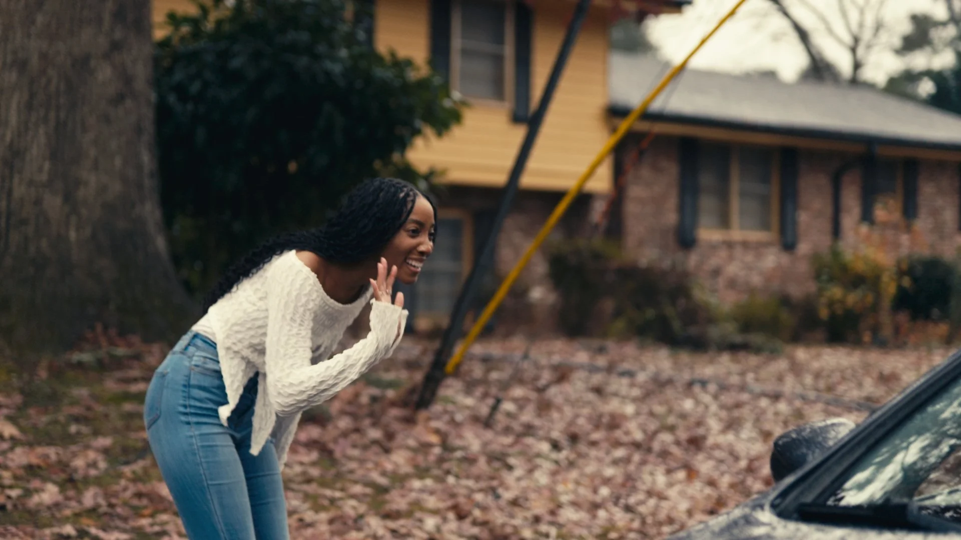 A young woman with long black hair, wearing a white sweater and blue jeans, is smiling and talking on her cell phone while leaning towards a car that appears to have stopped on the side of the road, with fallen autumn leaves on the ground and a house in the background.