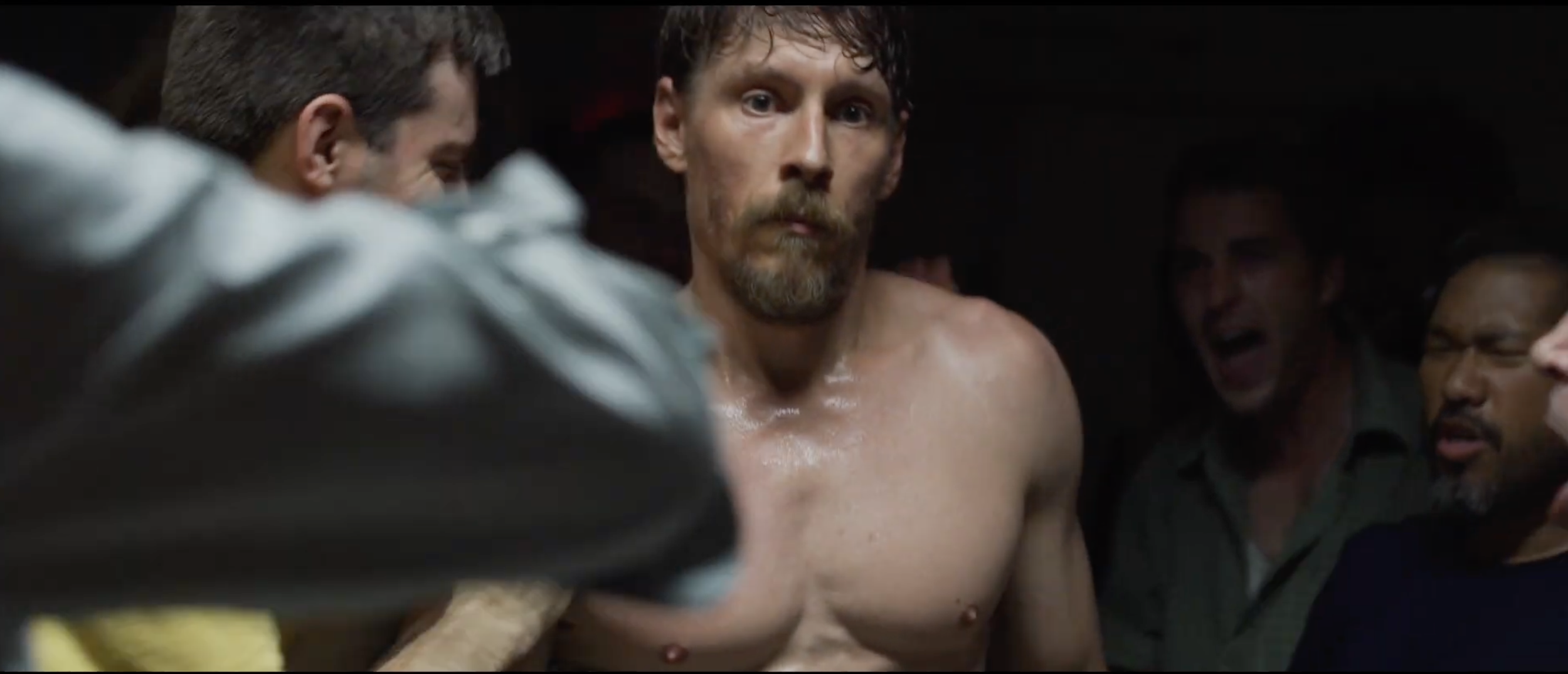A shirtless male boxer with a beard and curly hair in a boxing locker room surrounded by men, some of whom appear to be coaches or trainers, in a tense moment before a match.
