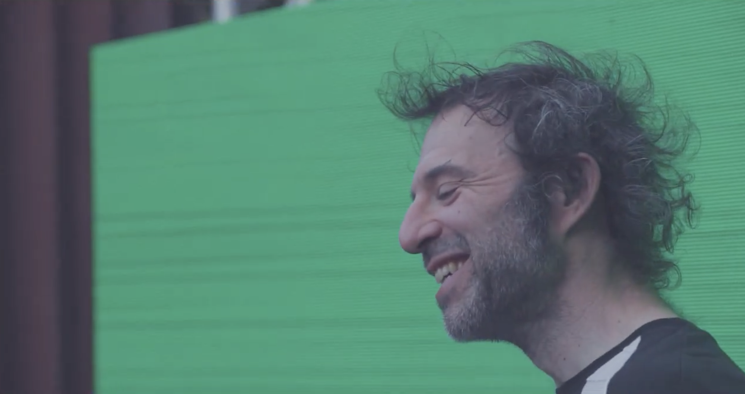 Close-up of a smiling man with curly hair and a beard, standing in front of a green background.