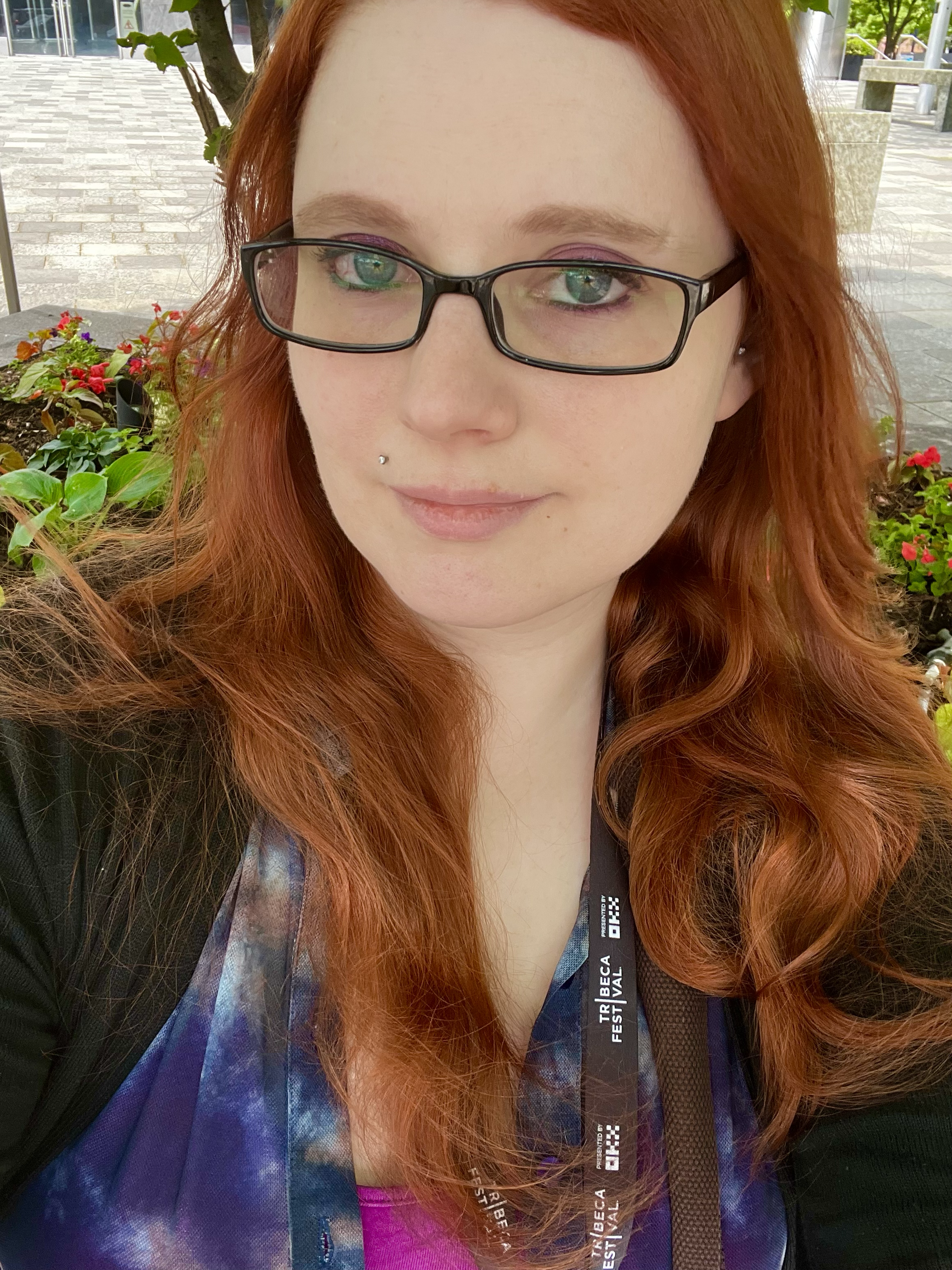 Close-up of a woman with long red hair, wearing glasses, a pink shirt, and a lanyard, outdoors near trees and flowers.