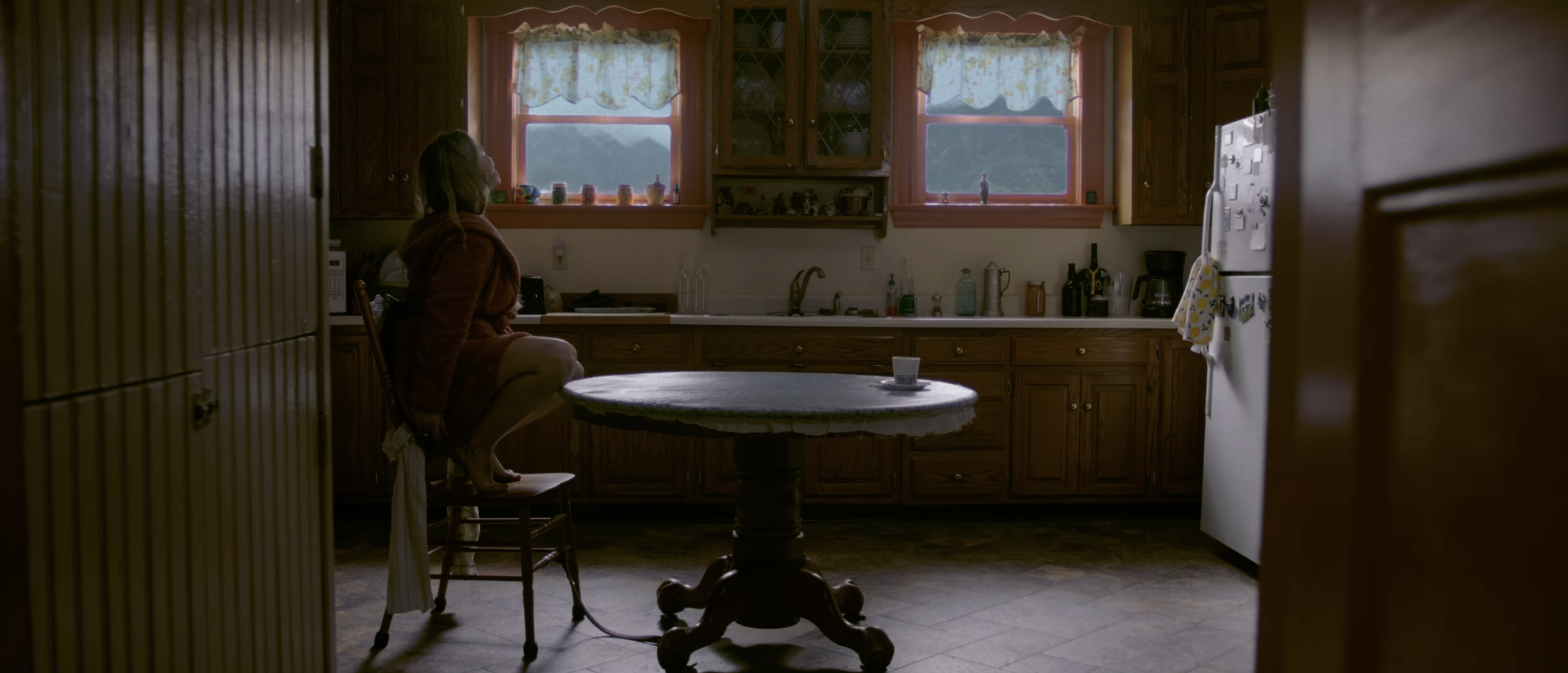 A woman sitting on a chair in a kitchen, looking out the window with a peaceful expression. The kitchen has wooden cabinets, a white refrigerator, and various kitchen appliances on the countertops. There is a round table in the center with a mug on it, and the window has floral curtains.