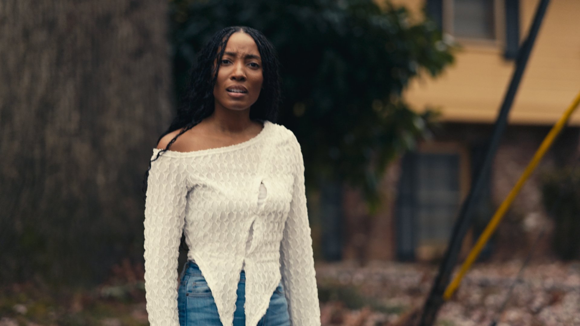 A woman with dreadlocks and a worried expression stands outdoors near a tree, wearing a white, distressed sweater and blue jeans.