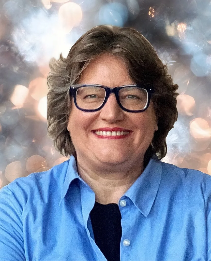 A smiling person with short, wavy hair and glasses, wearing a blue collared shirt, with a blurred background of multicolored bokeh lights.