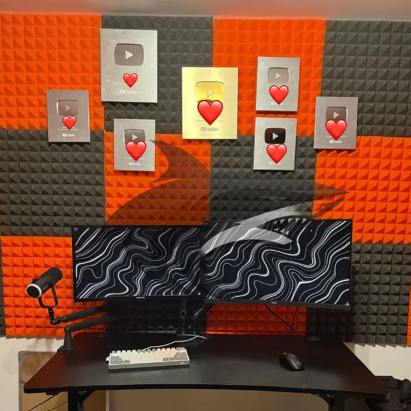 A recording or streaming studio with two computer monitors on a black desk, a keyboard, and a mouse. The wall behind features large YouTube play button awards with some Heart emojis on a background of orange and black acoustic foam panels.
