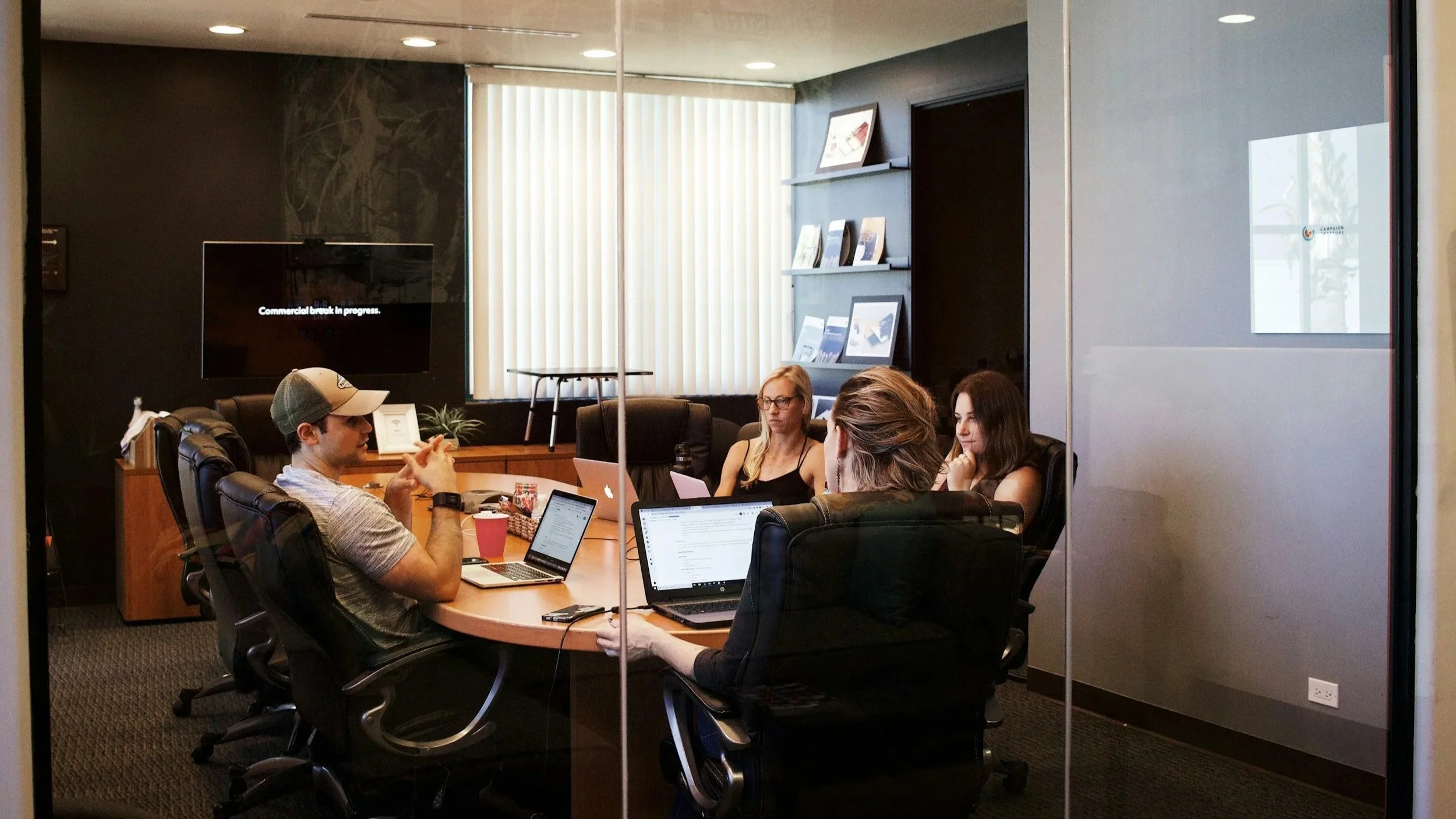 Four people sitting around a conference table in a modern office, working on laptops and tablets, with a large black screen on the wall displaying "Commercial break in progress."