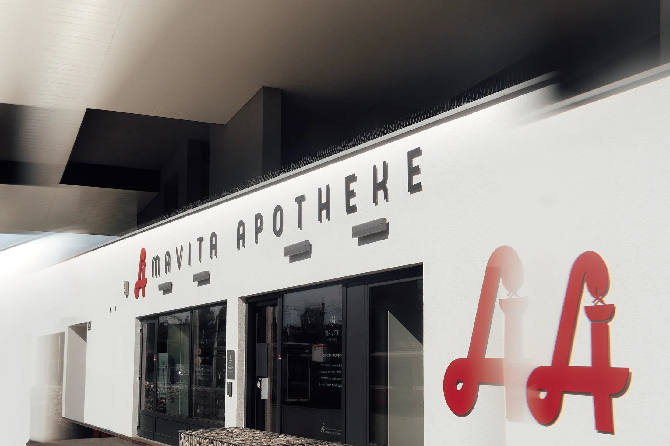 Außenansicht eines medizinischen Fachgeschäfts mit weißen Wänden, schwarzen Fenstern und Eingangstüren, das den Namen 'Mayvita Apotheke' trägt. Es gibt außerdem große rote und graue Schriftzüge und Symbole an der Fassade.