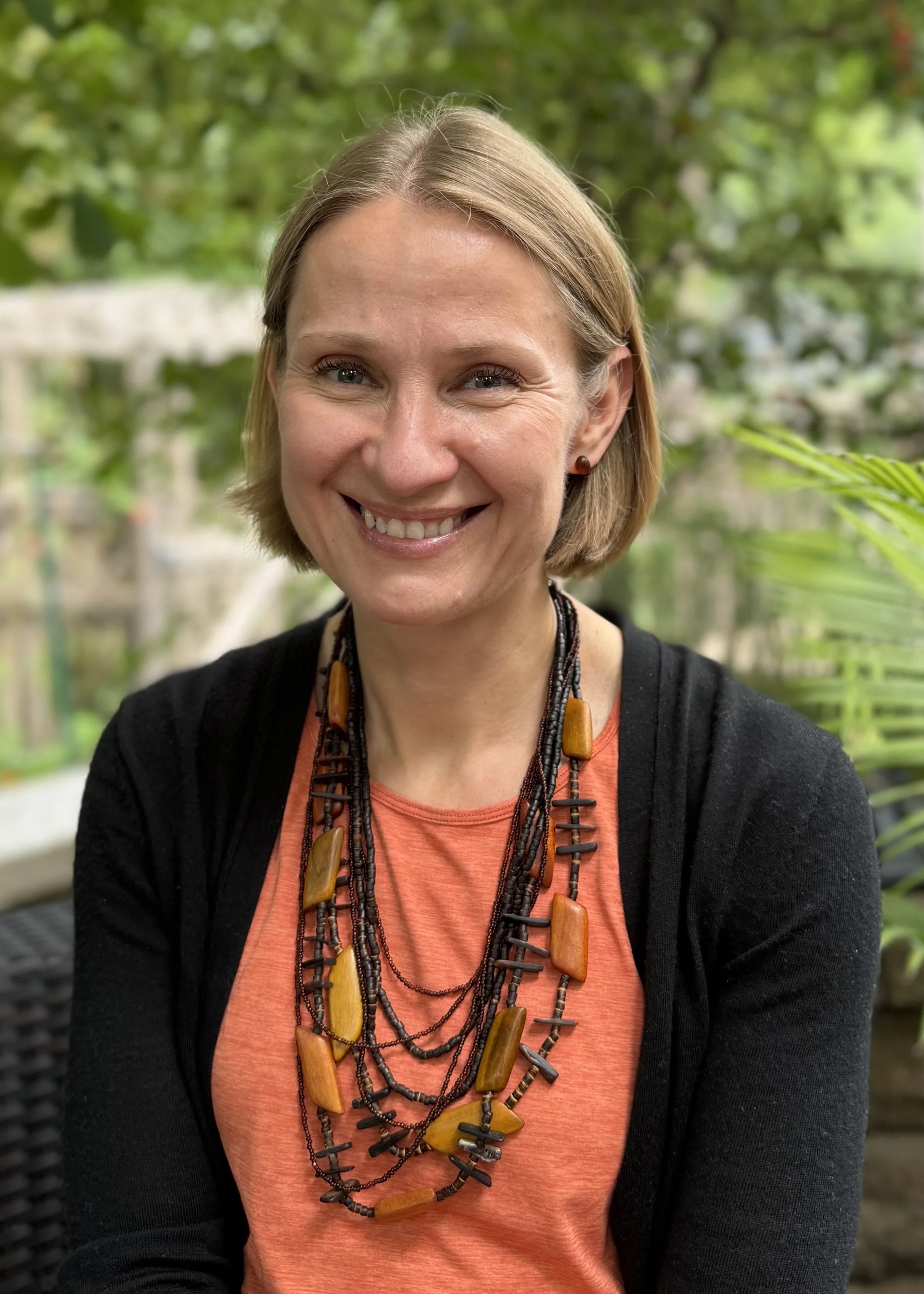 A woman with short blonde hair smiling, wearing an orange shirt, black jacket, and multiple beaded necklaces, outdoors with green foliage in the background.