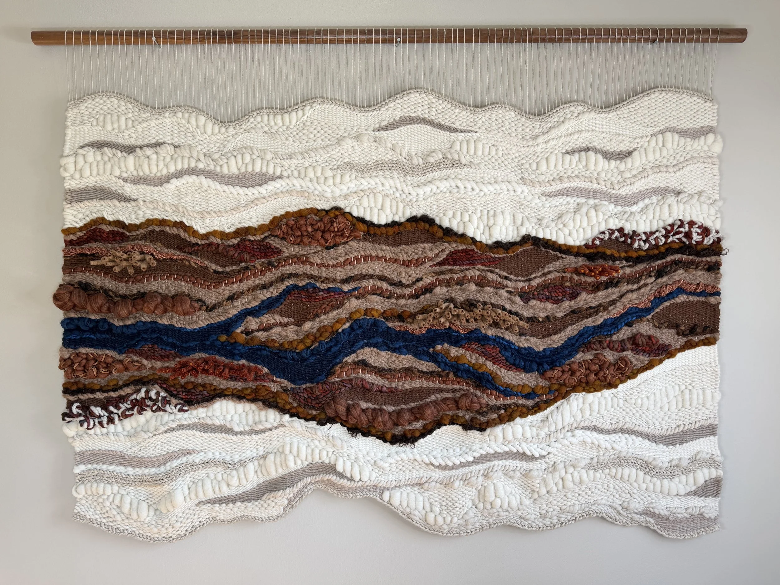A textile wall hanging with layered wavy patterns made from various yarns and fabrics, mounted on a wooden rod.