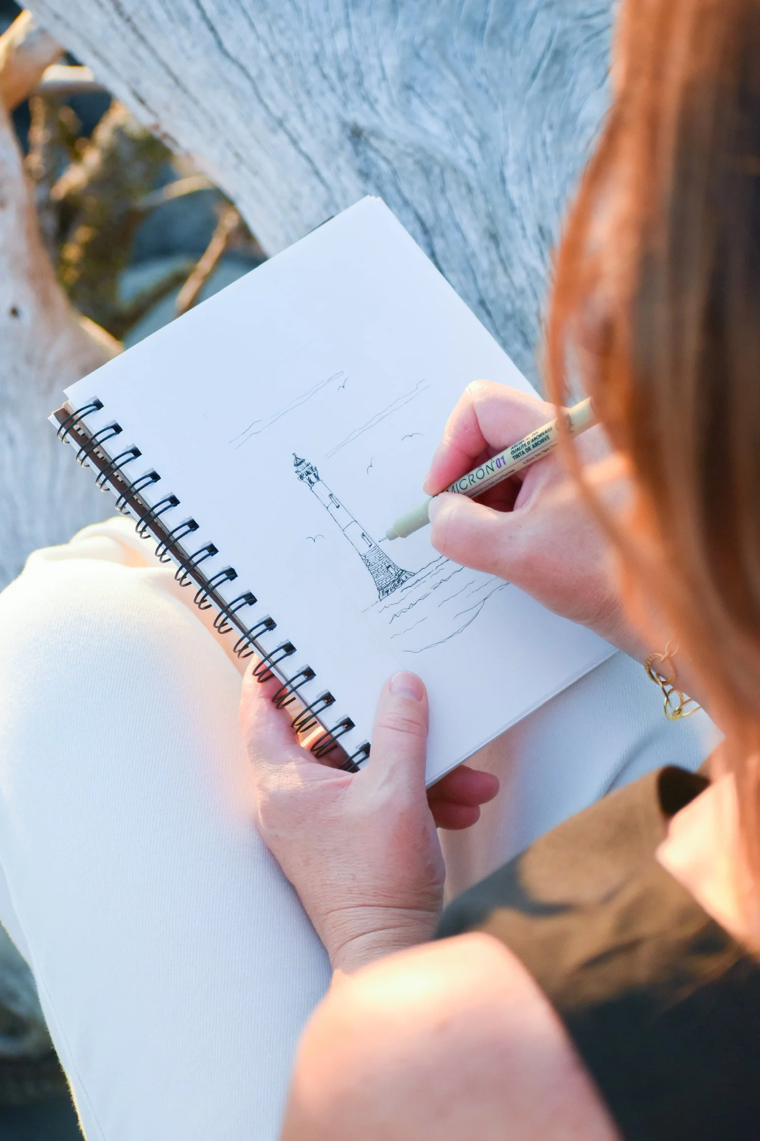Person sitting outdoors, drawing a lighthouse on a spiral notebook, with their right hand holding a pen and wearing a light bracelet. The background includes blurry tree branches and a wooden surface.