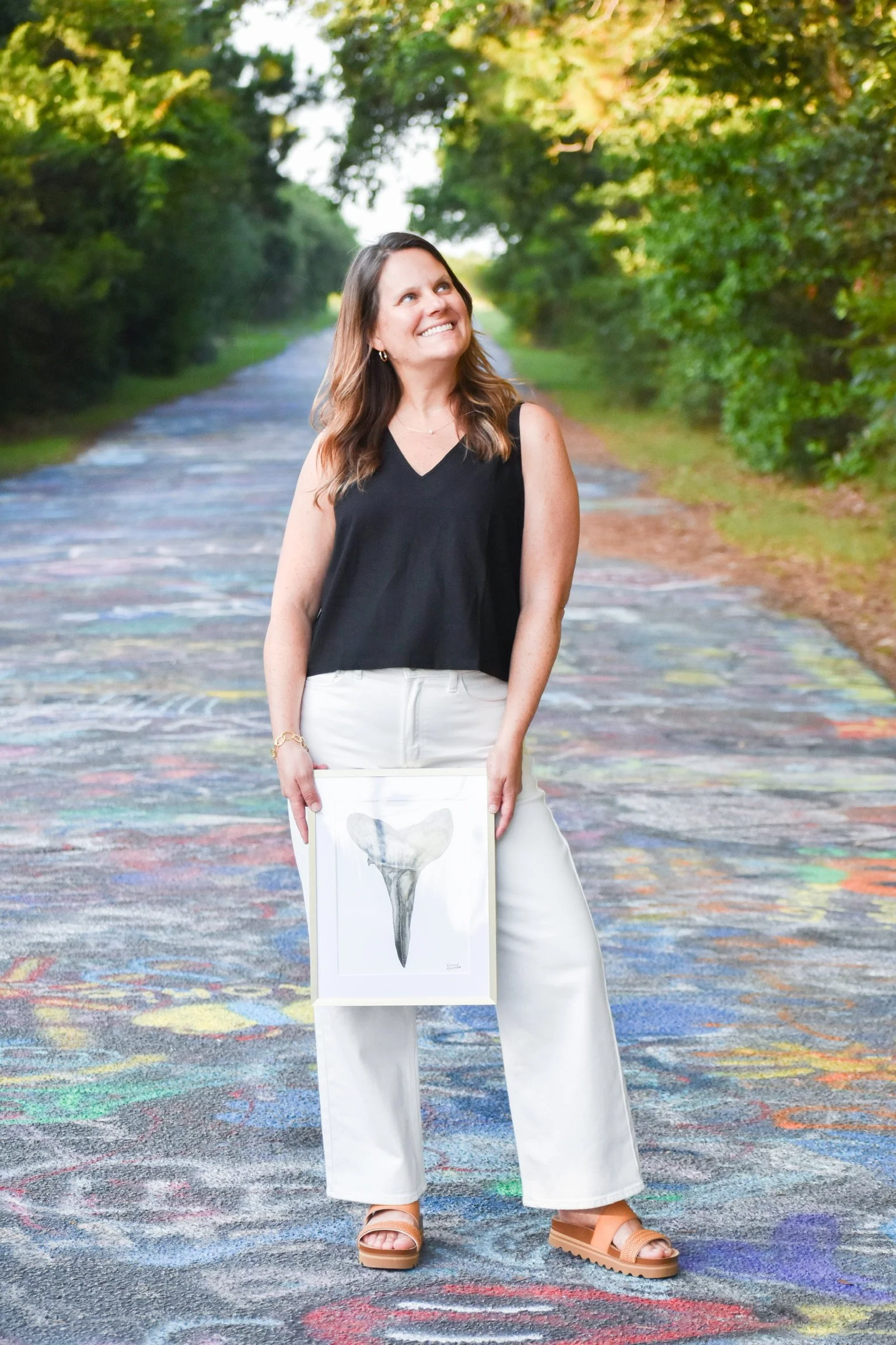 A woman in white pants and beige sandals holding a framed bird skull artwork, standing on a colorful outdoor carpet with green trees in the background.