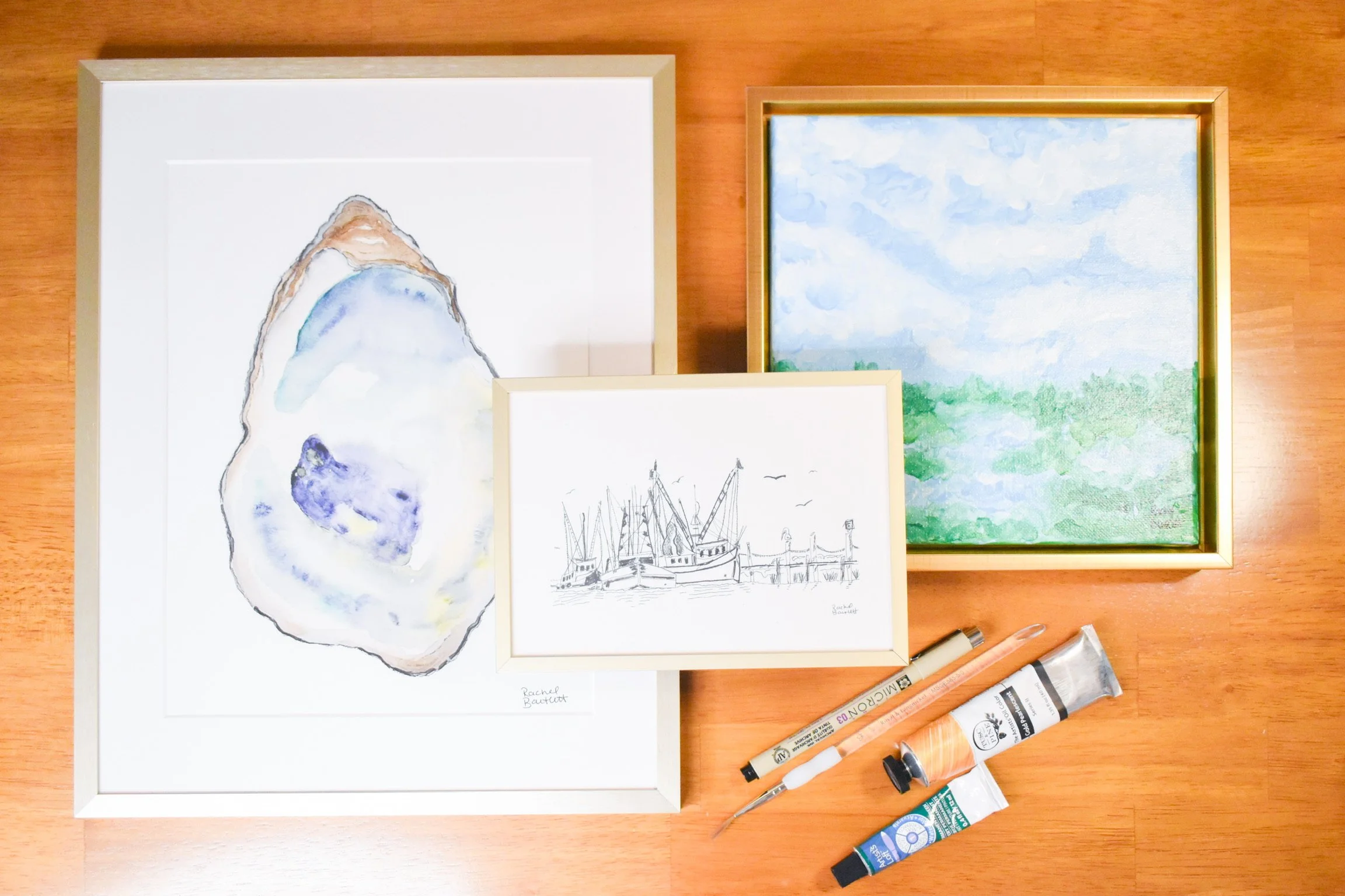 A collection of watercolor paintings, sketches, and art supplies on a wooden surface, including a mountain scene, a harbor with boats, and a landscape with a cloudy sky.