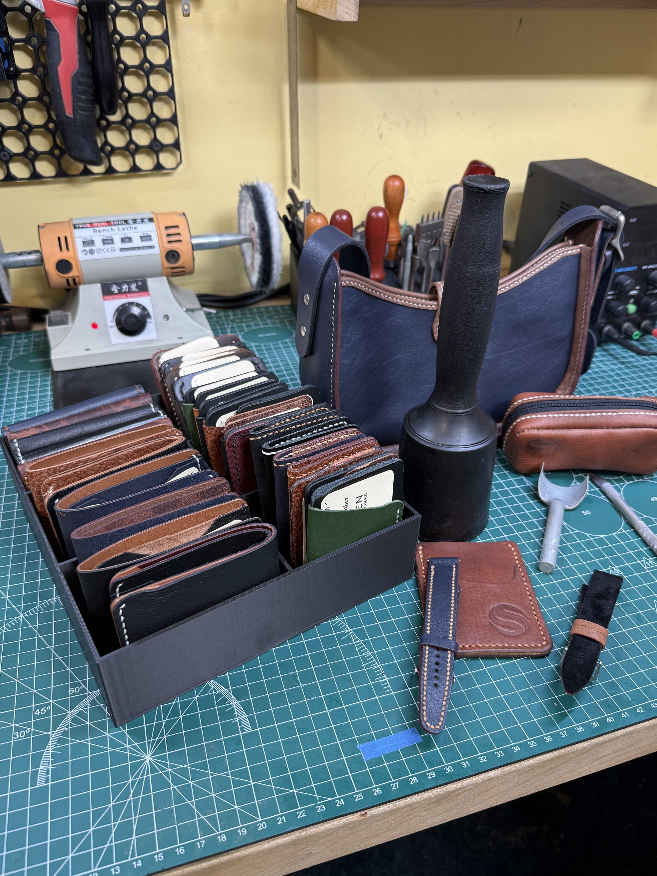 Leatherworking workbench with leather swatches, tools, and equipment including a leather edge beveler, cutting tools, a leather strap, and a belt on a green cutting mat.