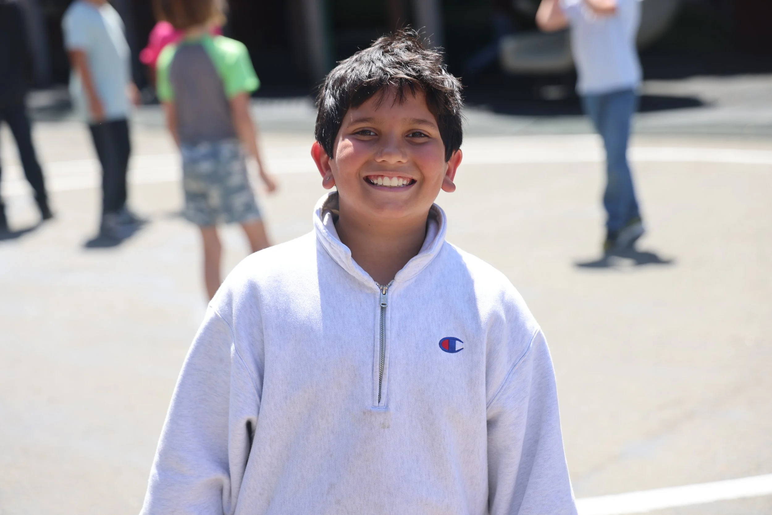A smiling boy with short dark hair and earrings, wearing a light gray hoodie, standing outdoors with a blurred background of other children and a black vehicle.