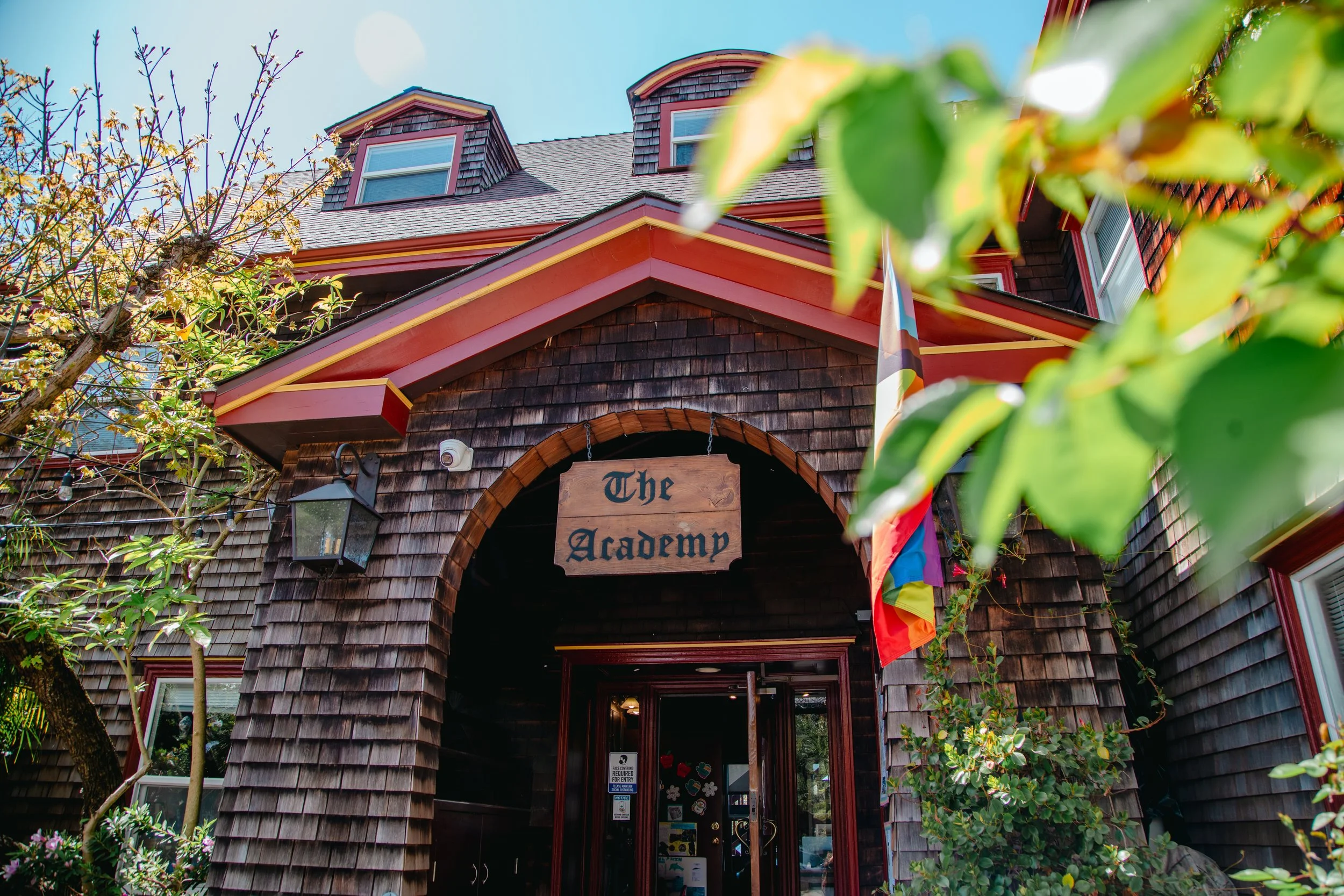 The entrance of a building with a sign that says 'The Academy'. The building has a wood-shingled facade, a red trim, and three dormer windows on the roof. There is a rainbow flag hanging next to the entrance, and the door has decorative stickers on it. Trees and greenery surround the building.