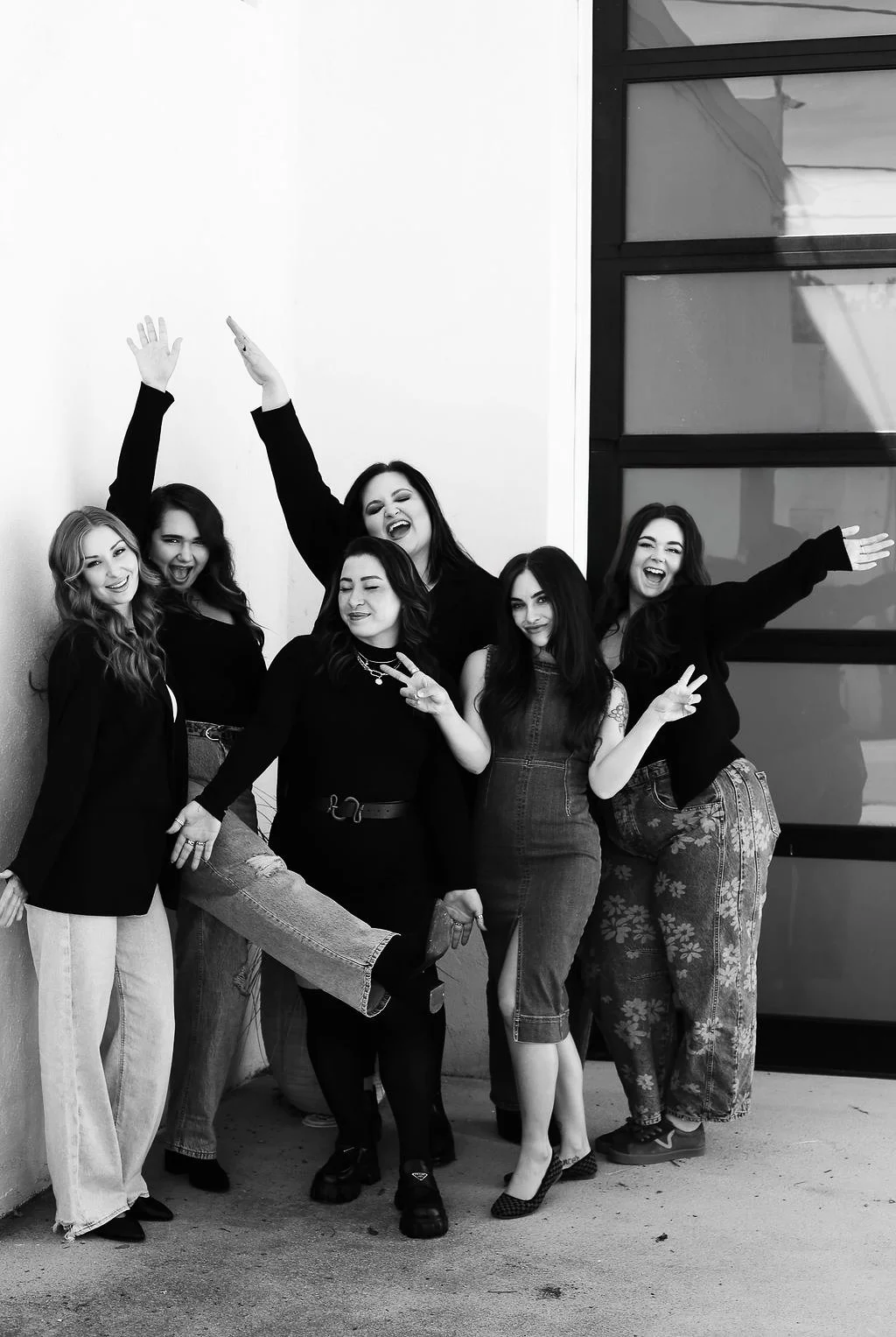 A group of six Emily Anne Esthetics employees posing happily outside a building, making peace signs, with some raising their arms and laughing.