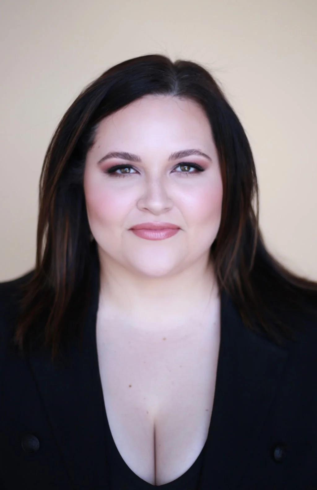 A woman with long dark hair, makeup, and a black top, smiling slightly against a neutral background.
