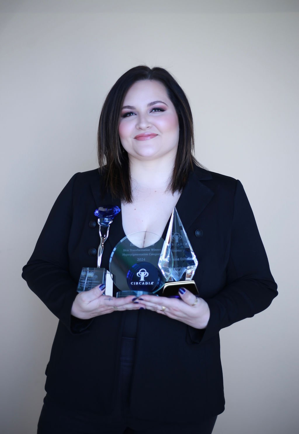 A woman with dark hair and makeup holding multiple award trophies, dressed in a black blazer, smiling in front of a plain beige background.