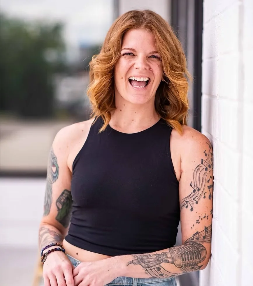 A woman with red hair and tattoos laughing while leaning against a white wall.
