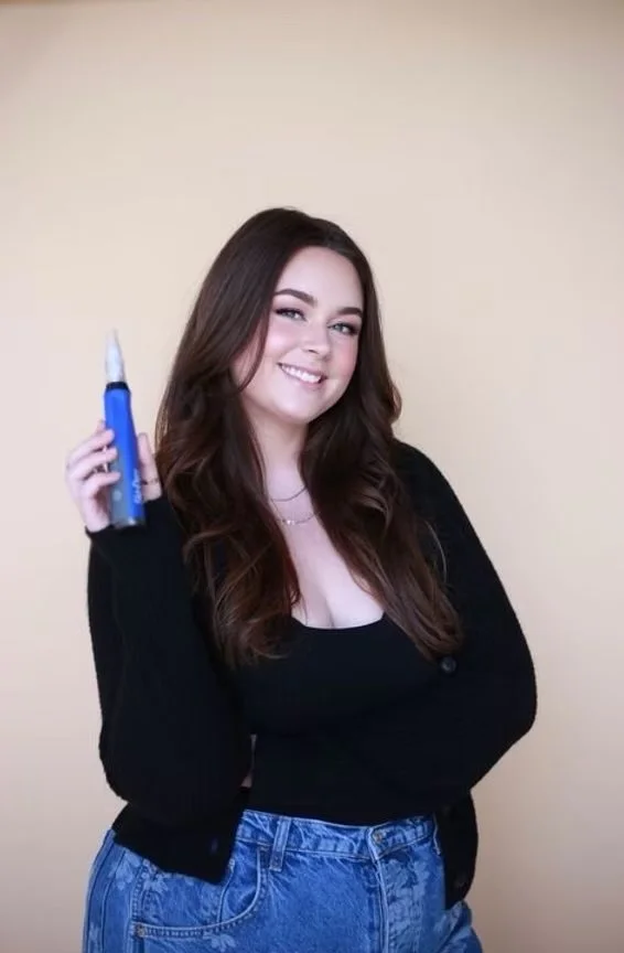 Smiling young woman with long brown hair holding a SkinPen, standing against a beige background.