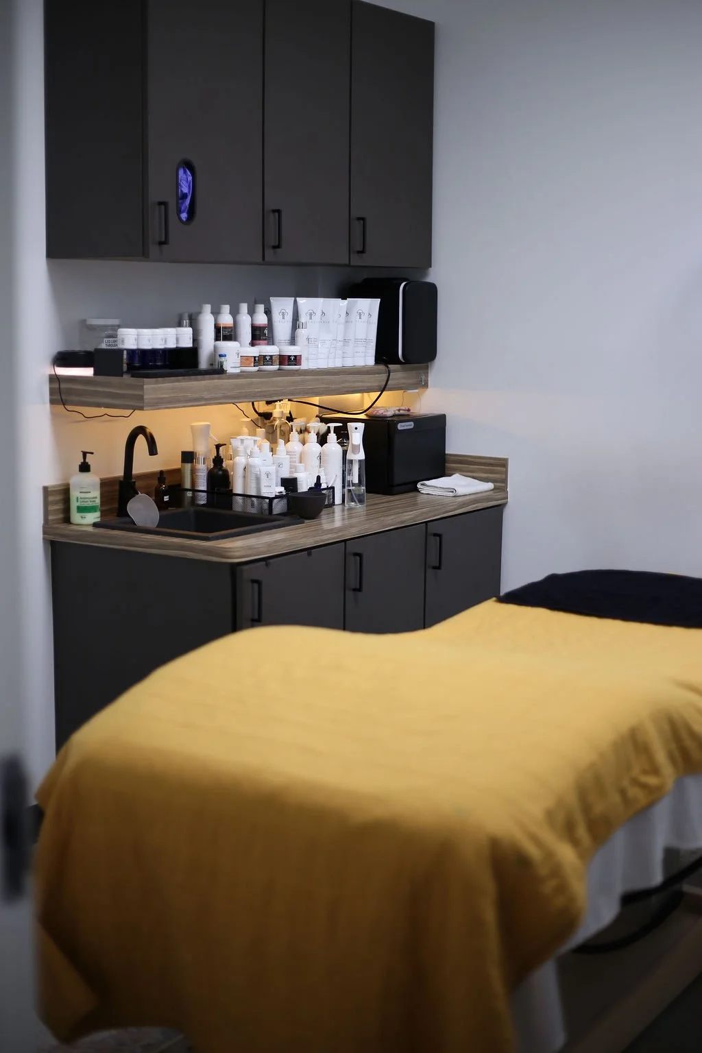 A medical or massage therapy room with a treatment bed covered in a yellow sheet, a small sink area with various bottles and skincare products, and cabinets above and below the counter.