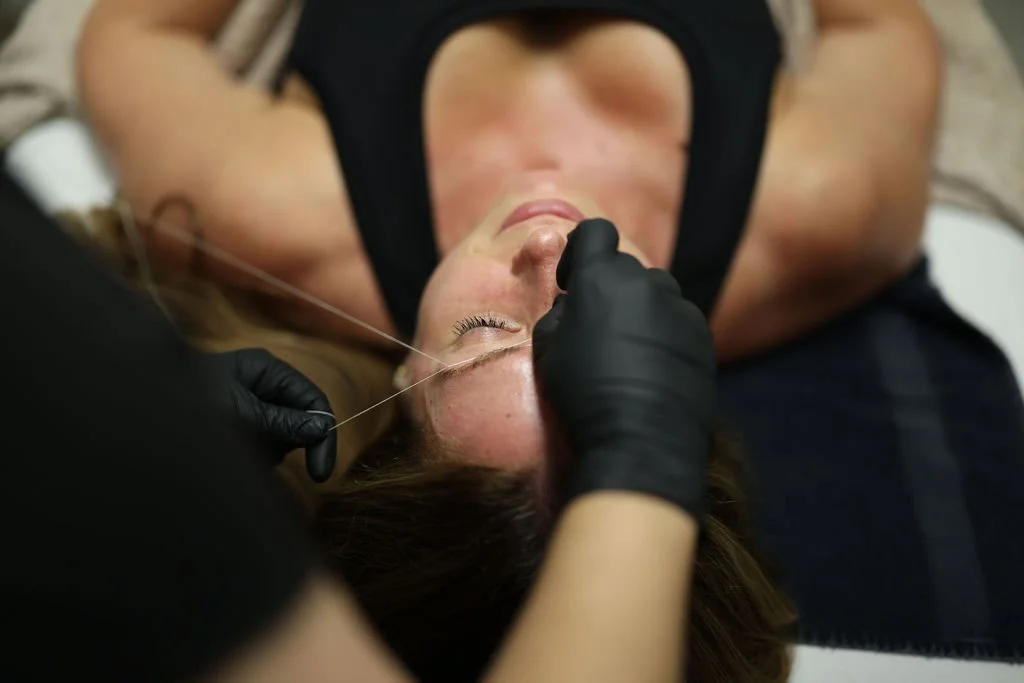 A woman receiving brow threading, lying on a reclined chair while a technician wearing black gloves performs the procedure.