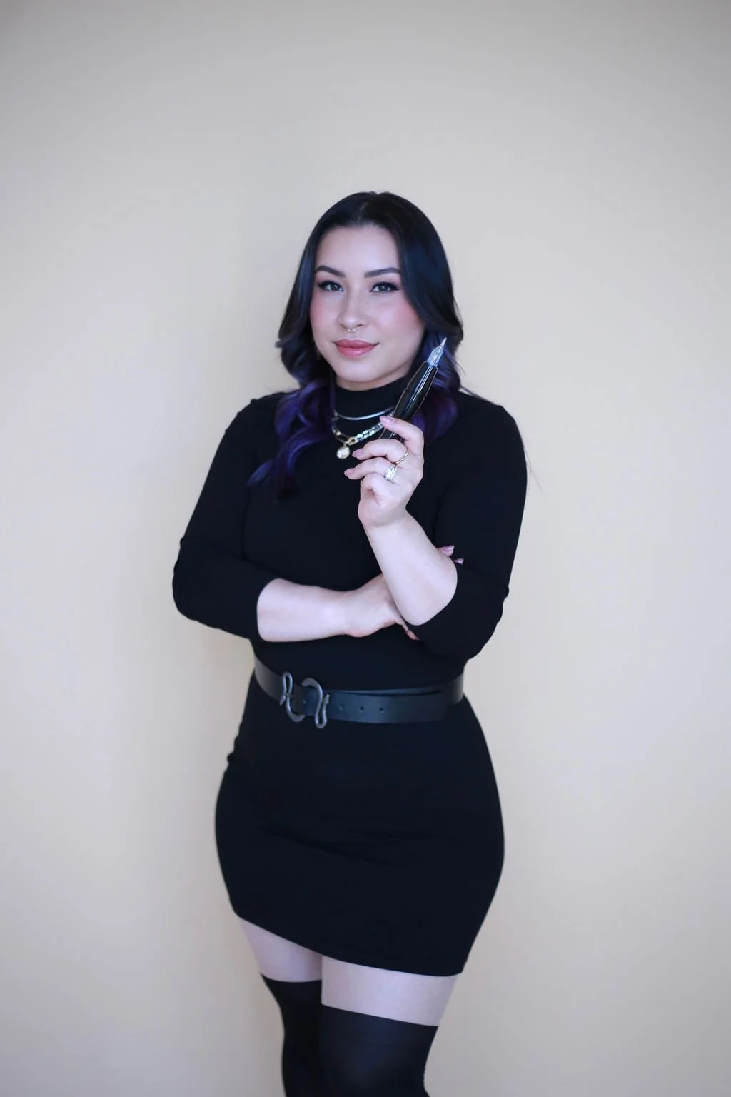 A woman with black and purple hair wearing a black dress, holding a permanent makeup tool, standing against a plain light background.