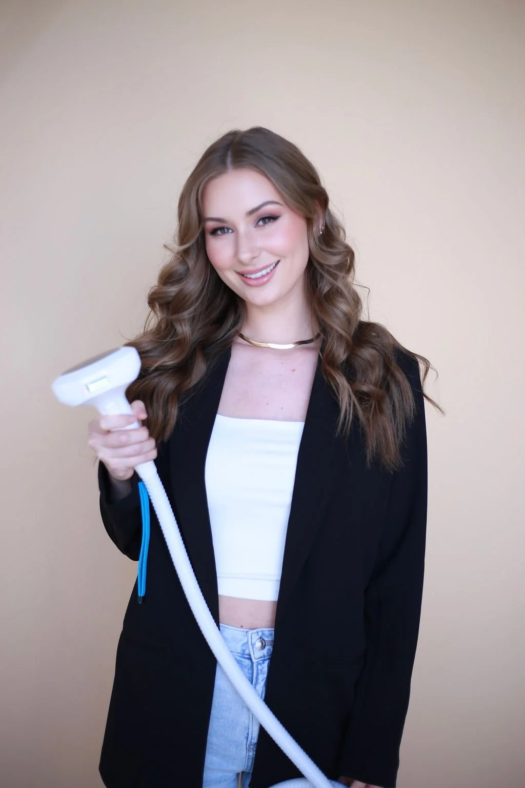 A young woman with long wavy brown hair smiling and holding an aesthetic tool.