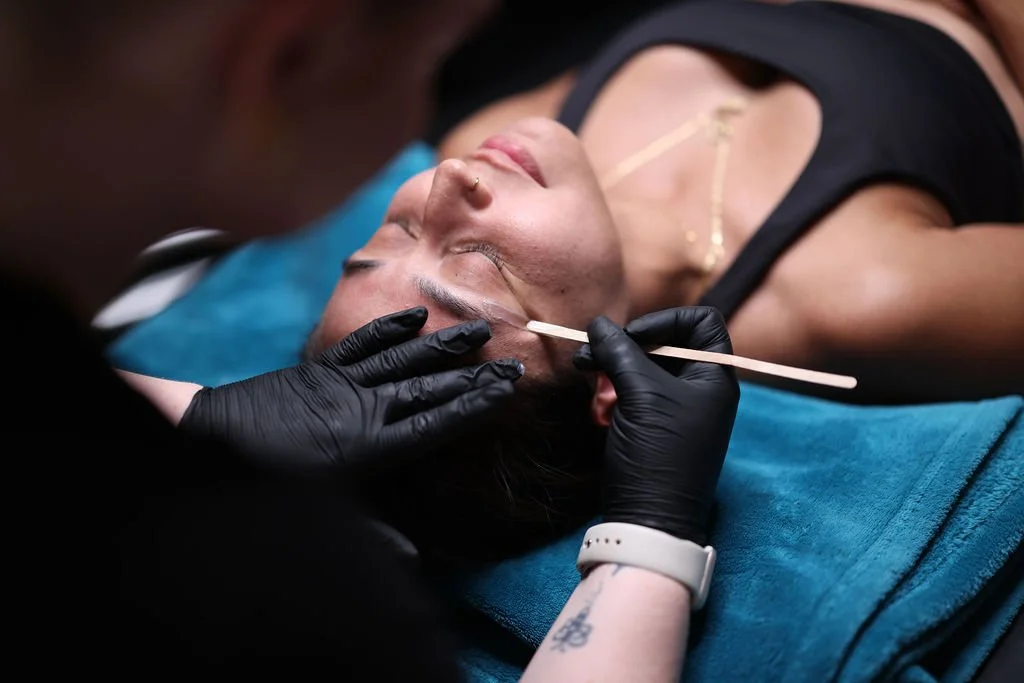 A woman lying down on her back with her eyes closed, receiving a brow lamination treatment from technician.
