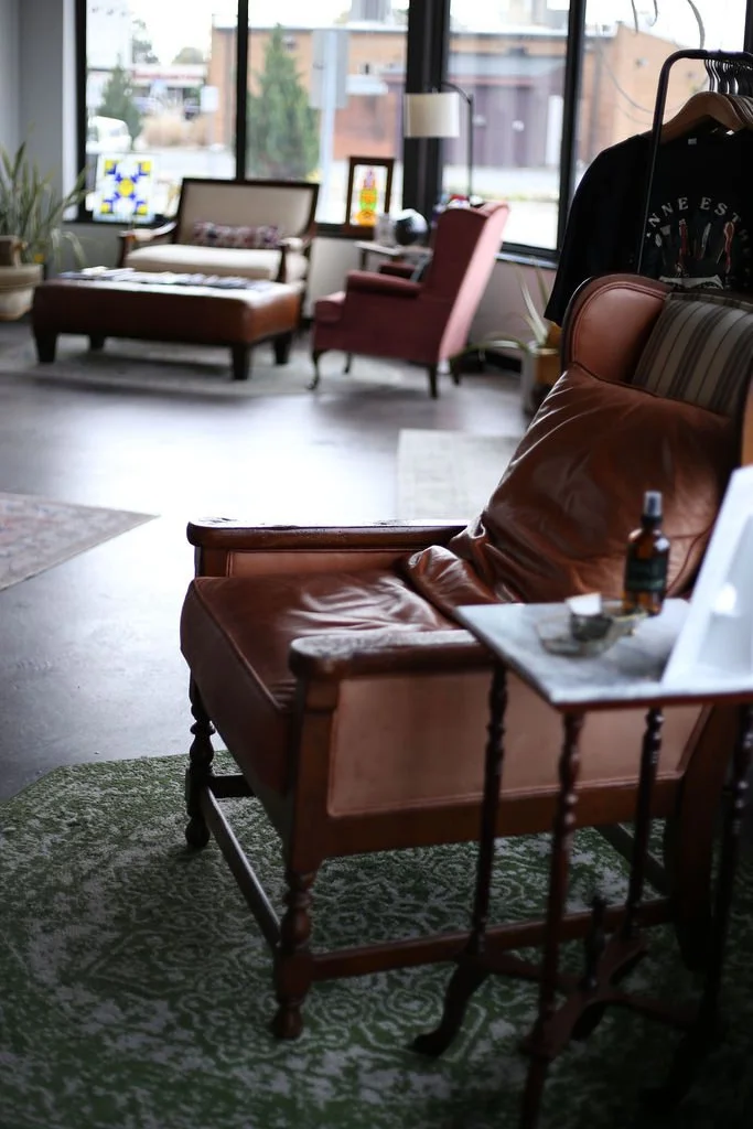 Cozy vintage living room with leather armchair, side table, sofa, armchair, large windows, plants, and decorative stained glass panels.
