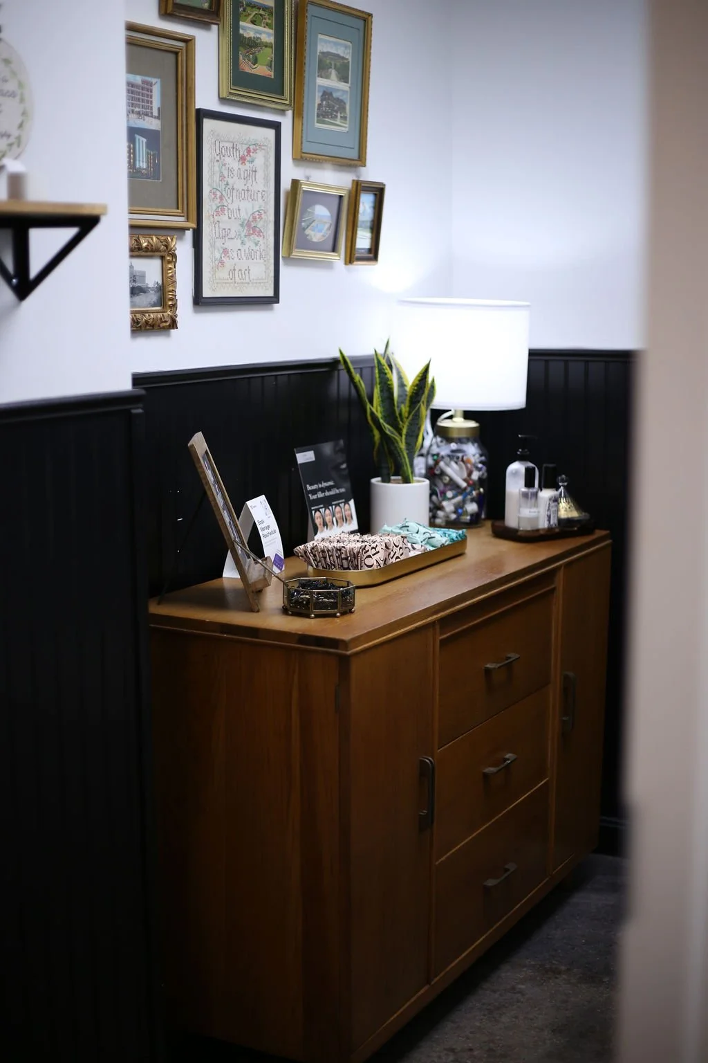 A wooden cabinet with three drawers and two doors, placed against a wall with black wainscoting. On top of the cabinet are a potted snake plant, a large white table lamp, a glass jar filled with small objects, a tray with tissues, and some lotion and soap dispensers. Above, a wall display features various framed pictures and art, including landscape photos and a cross-stitched quote. The setting appears to be a cozy, decorated corner of an interior room.