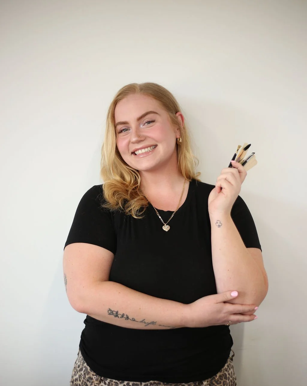 A smiling woman with blonde, shoulder-length hair, wearing a black shirt and leopard-print pants, holding makeup brushes in her right hand, standing against a plain white wall.